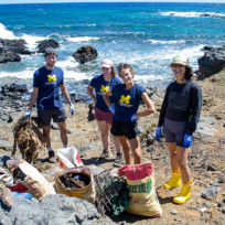 A group of people stands on a rocky coastline with collected debris and sacks. Waves crash in the background. They are wearing casual outdoor clothing and gloves.