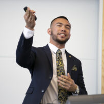 A person in a suit and paisley tie gestures while speaking in front of a whiteboard.