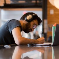 Man wearing headphones and glasses writing in a notebook at a table with a laptop and water bottle, indoors.