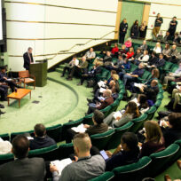 A speaker addresses an audience in a tiered lecture hall. Panelists sit beside the podium while attendees, seated in green chairs, listen and take notes.