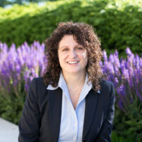A person with curly brown hair is smiling, wearing a black blazer and light shirt. They are sitting outdoors with purple flowers and greenery in the background.