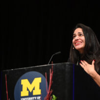 A woman stands at a podium with a University of Michigan logo, speaking and gesturing with one hand on her chest.