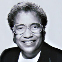 Portrait of a smiling woman wearing large glasses, a dark jacket, and a light blouse. The background is plain white.