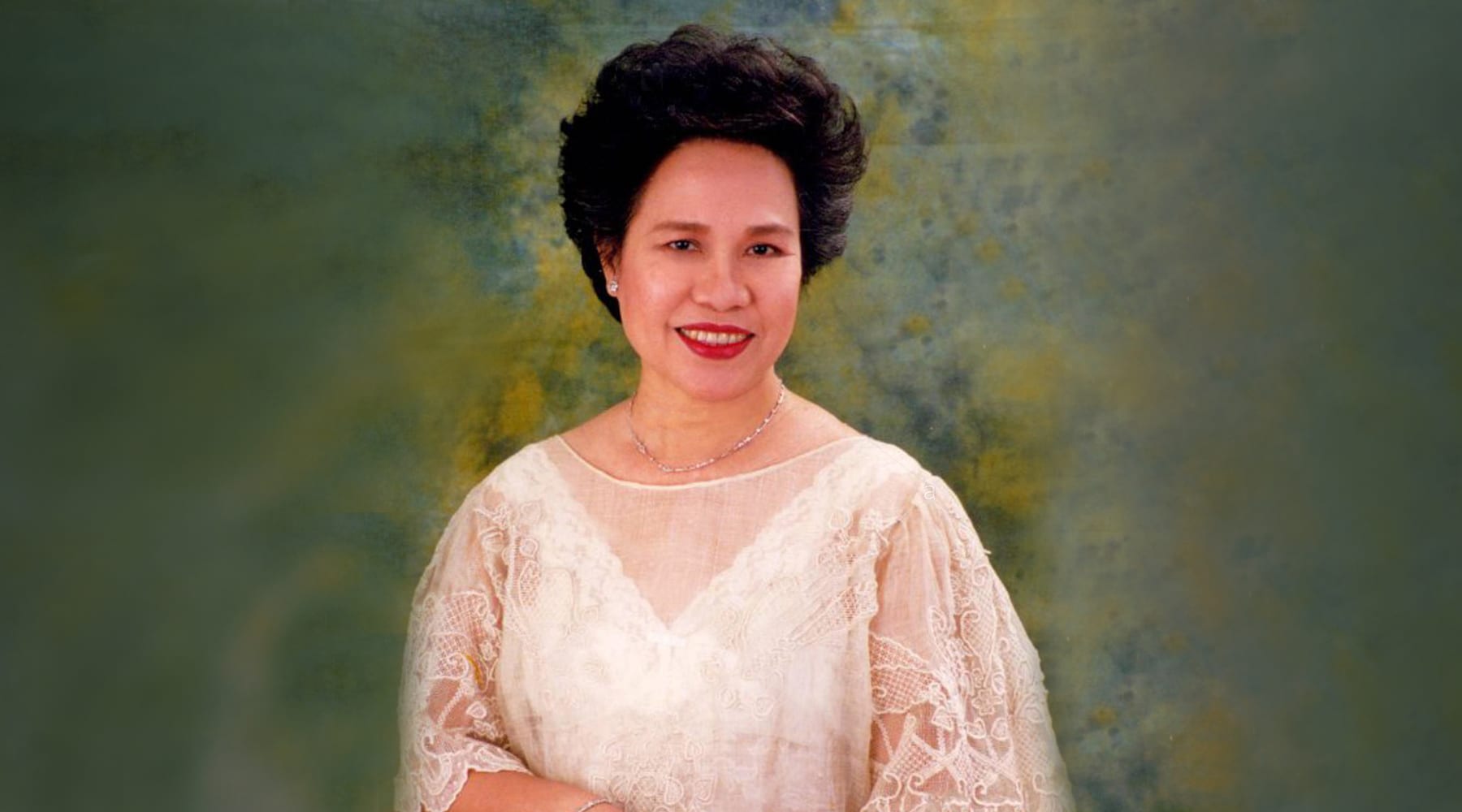 A woman in a cream lace dress poses against a green marbled backdrop, smiling with hands clasped.