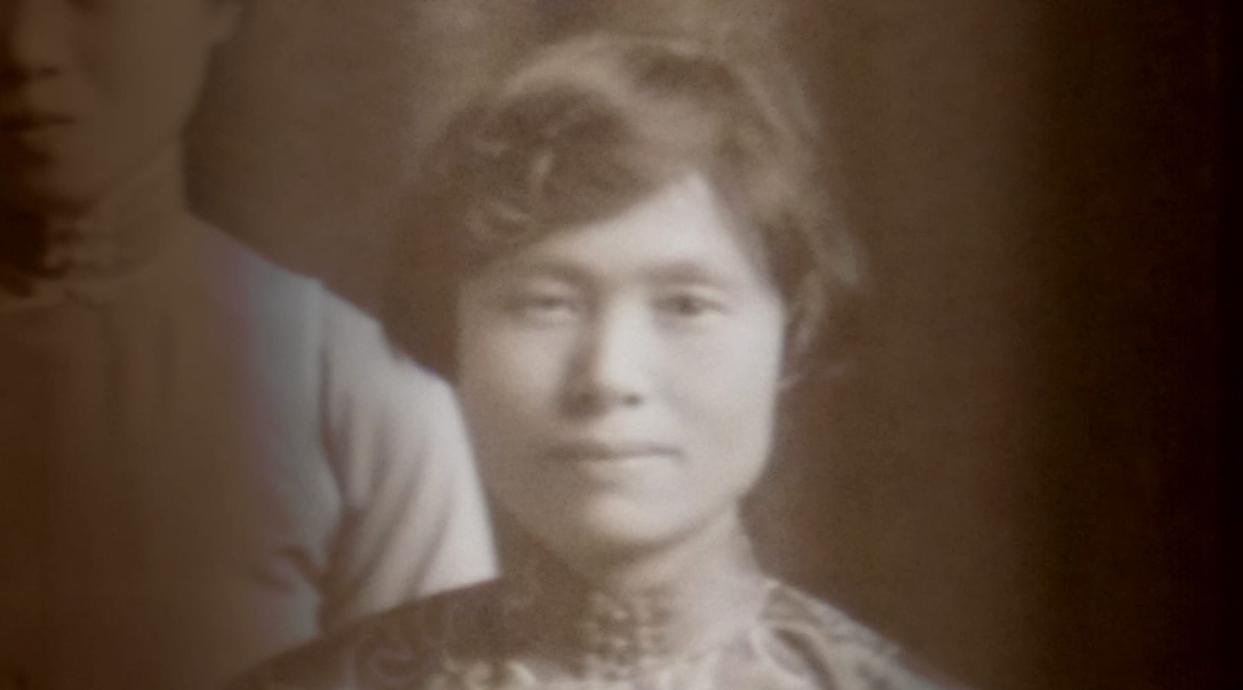 A sepia-toned vintage portrait of a woman with short hair, wearing a patterned garment, looking directly at the camera with a neutral expression.
