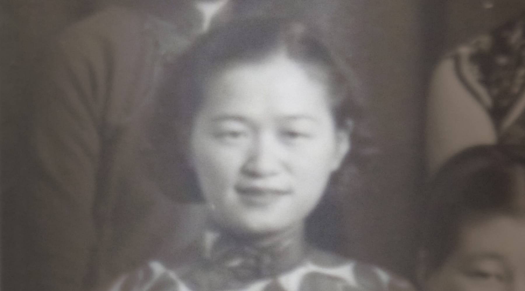 Black and white portrait of a woman, wearing a patterned top, looking at the camera, with other partially visible people in the background.