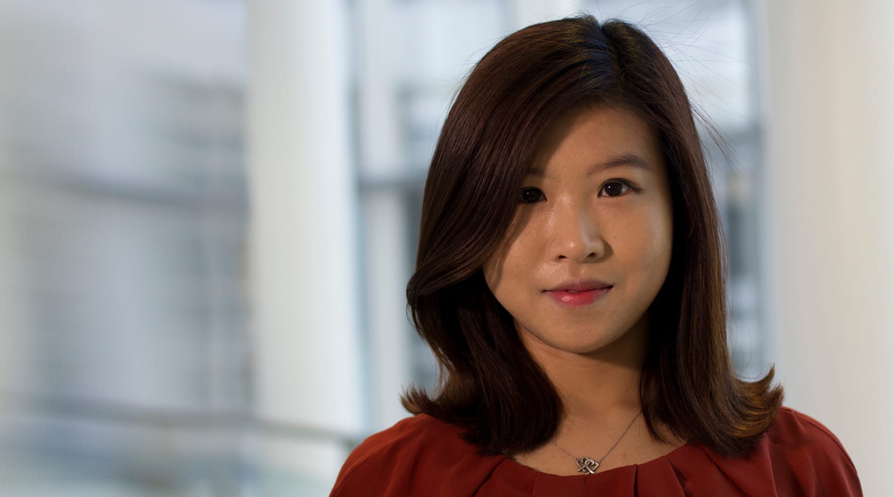 A person with shoulder-length brown hair is wearing a red top and a necklace, standing in front of a blurred background.