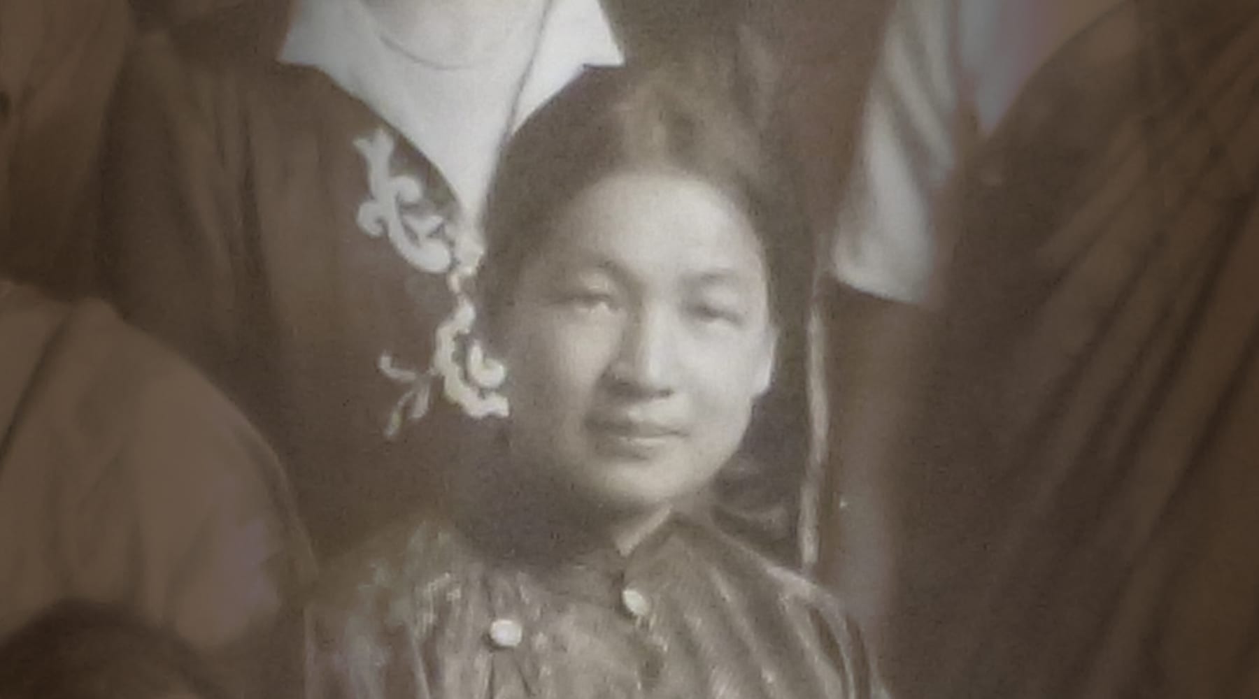 A black and white vintage portrait of a woman wearing traditional clothing, with an embroidered design visible on her attire. She is looking directly at the camera against a neutral background.