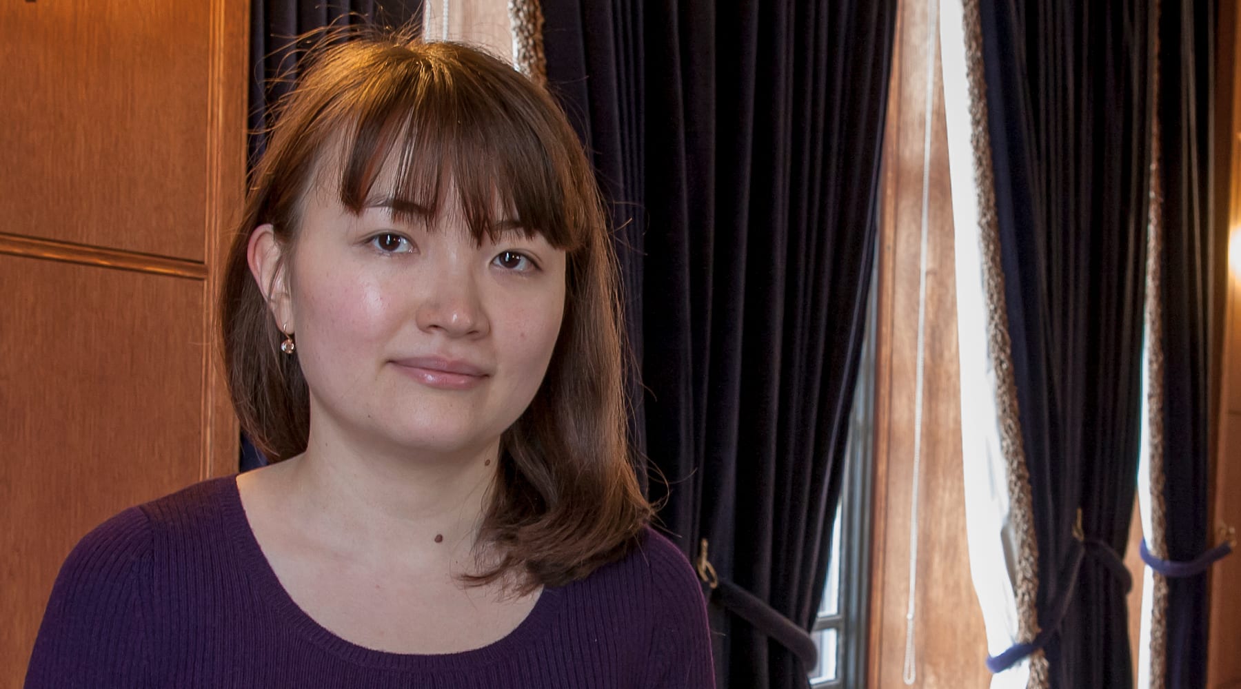 A woman with shoulder-length hair and a purple sweater stands indoors near dark curtains and wooden paneling.