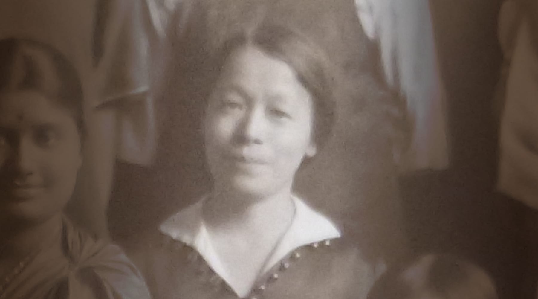 Black and white photo of a woman in a collared dress, seated and looking at the camera, with a neutral expression. Other people are partially visible around her.
