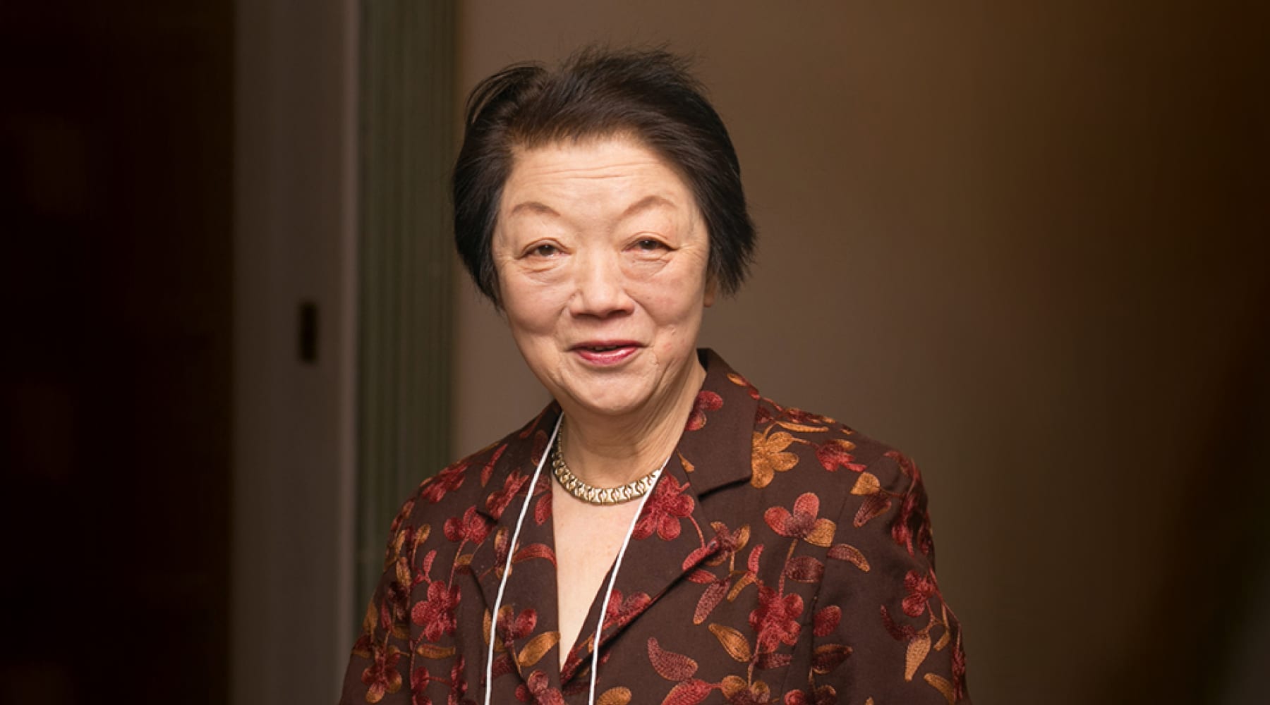 An older woman is wearing a floral patterned jacket and a necklace, standing against a neutral background.