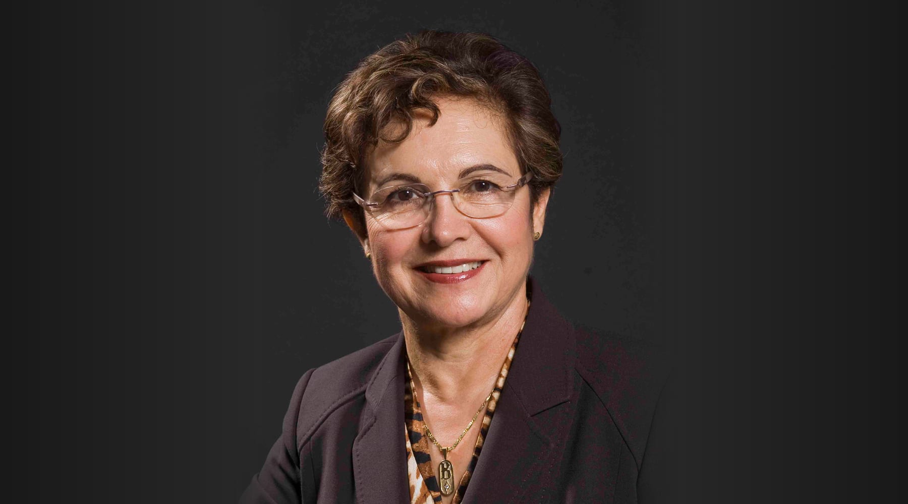 Woman wearing glasses and a dark blazer smiles against a dark background.
