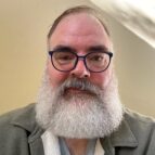 A person with a full white beard, glasses, and a green jacket faces the camera against a plain light-colored background.