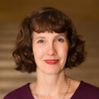 Woman with short curly brown hair and a necklace, wearing a maroon top, looking at the camera with a neutral expression against a blurred brown background.
