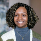 Woman with curly, shoulder-length hair and a warm smile, wearing a gray turtleneck, gold necklace with a cross, and a green and white sweater.