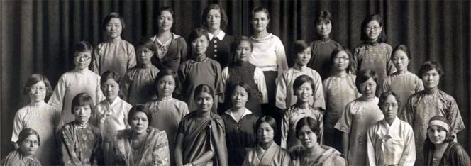 A black and white group photo of 26 women lined up in three rows, wearing a mix of traditional and early 20th-century Western attire, set against a dark curtain backdrop.