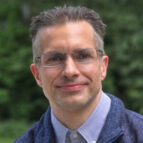 A person with short hair and glasses smiling, wearing a blue jacket over a collared shirt. Green foliage is in the background.