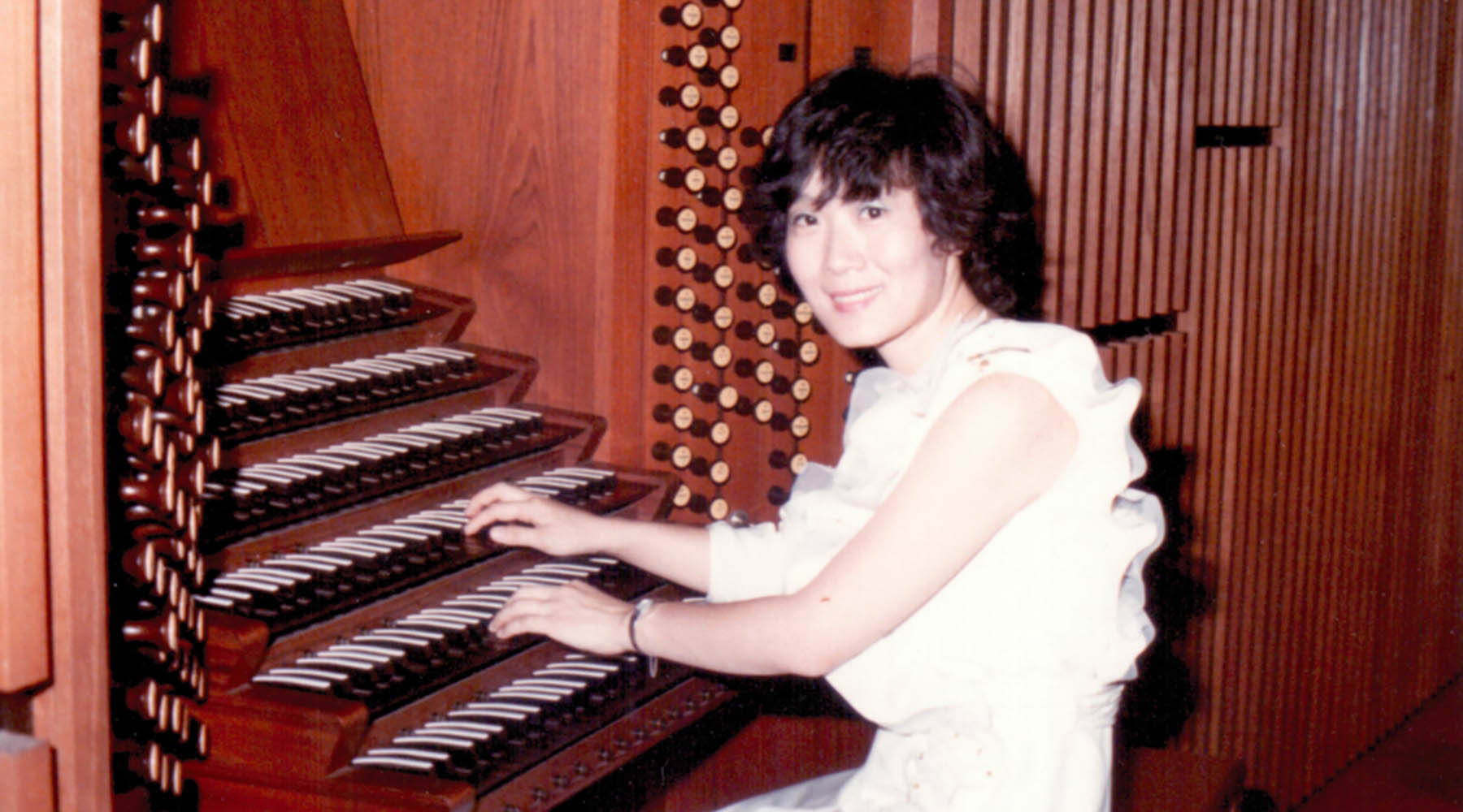 A person seated at a large organ, playing the keys, surrounded by numerous wooden pipes and stops, wearing a white outfit.