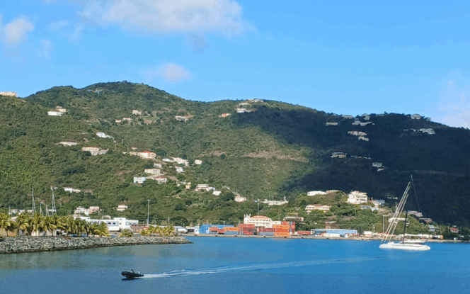 A coastal scene with green hills dotted with houses, colorful buildings near the shore, a sailboat, and a small motorboat on blue water under a clear sky.