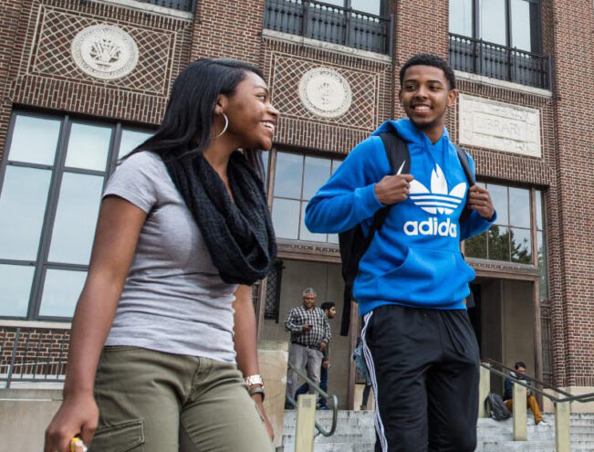 Two students walking down steps outside a brick building with large windows, one wearing a blue Adidas hoodie and the other holding a phone and smiling.