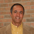 A man with short dark hair wearing a yellow shirt and light brown blazer stands in front of a brick wall, facing the camera and smiling slightly.