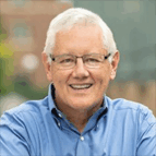 An older man with short white hair and glasses, wearing a blue button-down shirt, smiling outdoors with a blurred background.