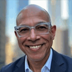 A bald man wearing clear glasses and a suit smiles at the camera with an urban background of blurred tall buildings.