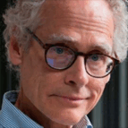 An older man with curly gray hair, glasses, and a striped shirt looks at the camera with a slight smile.