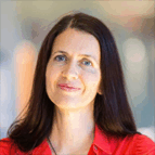 A woman with long dark hair, wearing a red shirt, is looking at the camera and smiling slightly against a blurred indoor background.