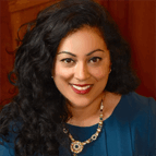 A woman with long curly black hair, wearing a blue top and a jeweled necklace, smiles while sitting in front of a wooden background.