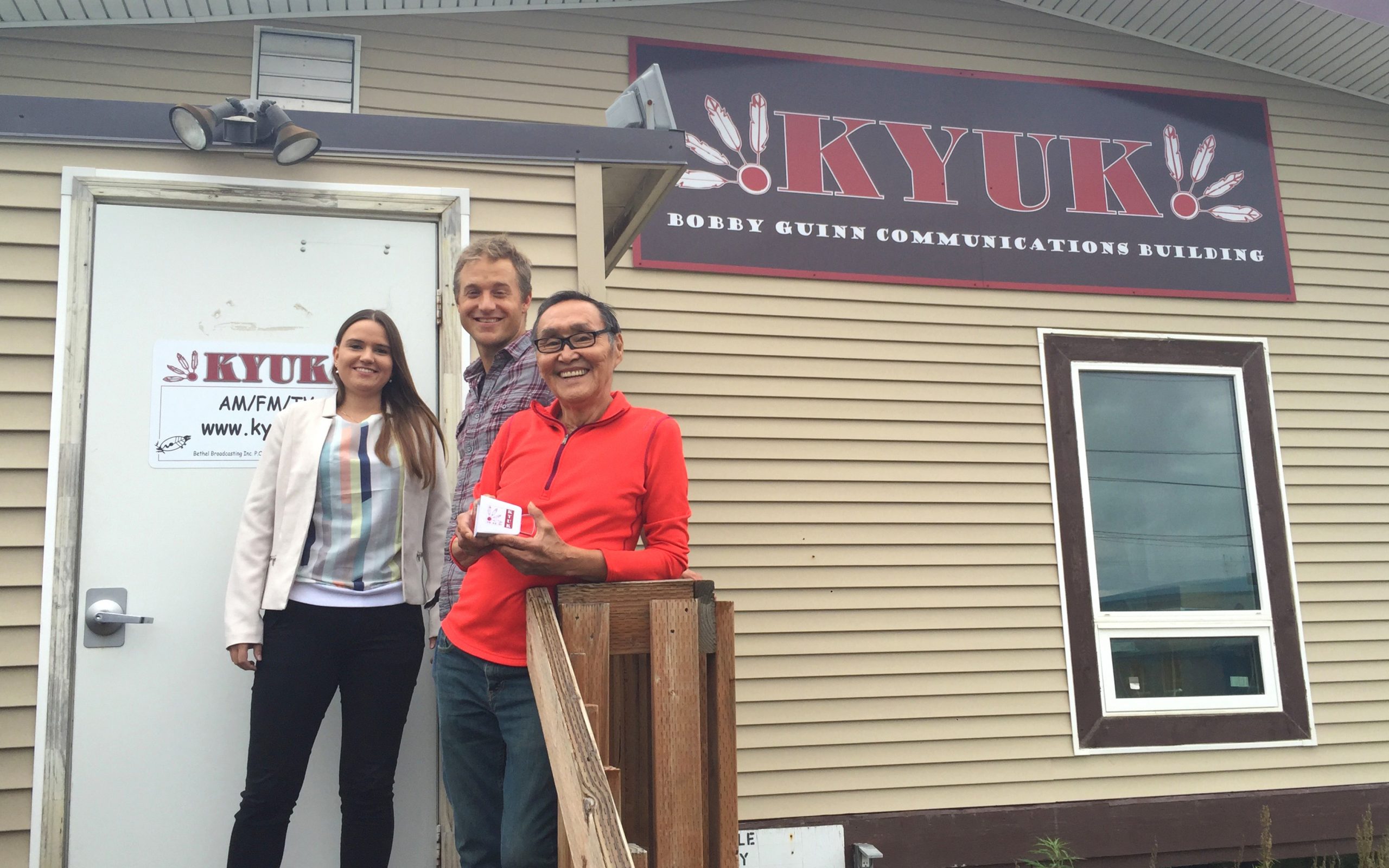 Three people stand smiling in front of the KYUK Bobby Gunn Communications Building, next to a sign with the station's details. One person holds a small box.