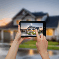 Person holding a tablet with smart home controls displayed, in front of a modern house at dusk.
