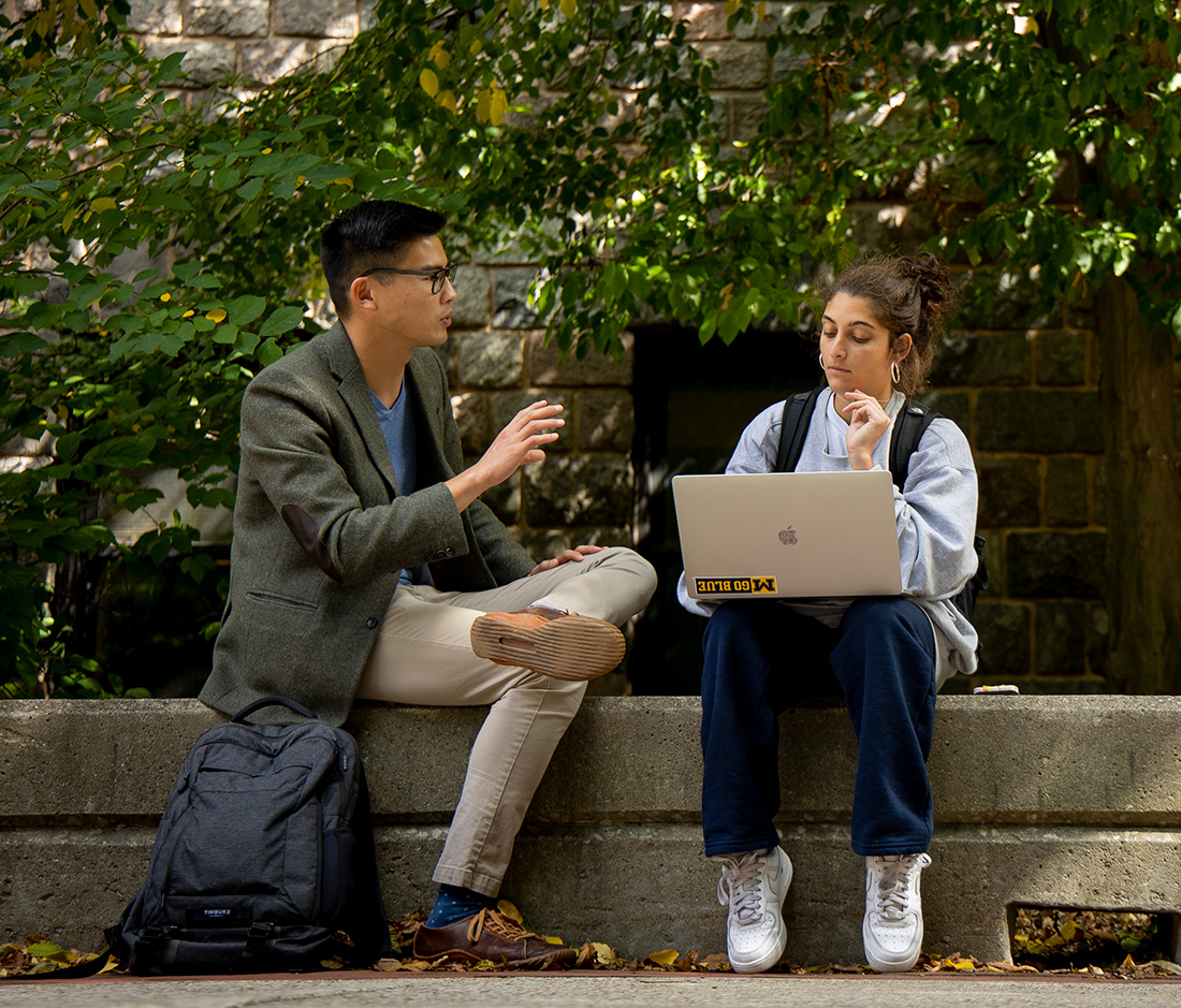 Two students sit outdoors on a stone ledge, engaged in conversation. One gestures while speaking, and the other listens, holding a laptop. Trees and a stone wall are in the background.