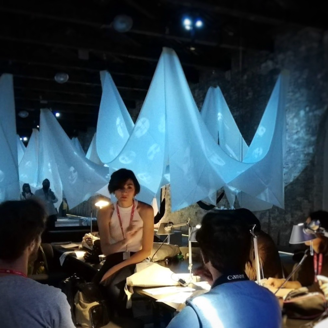 People sit and talk at tables in a dimly lit room with translucent fabric installations hanging from the ceiling, illuminated by blue light.