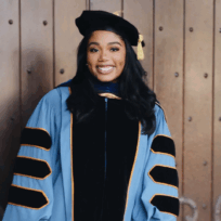 A woman in academic regalia with a doctoral gown and cap stands and smiles in front of a wooden door.