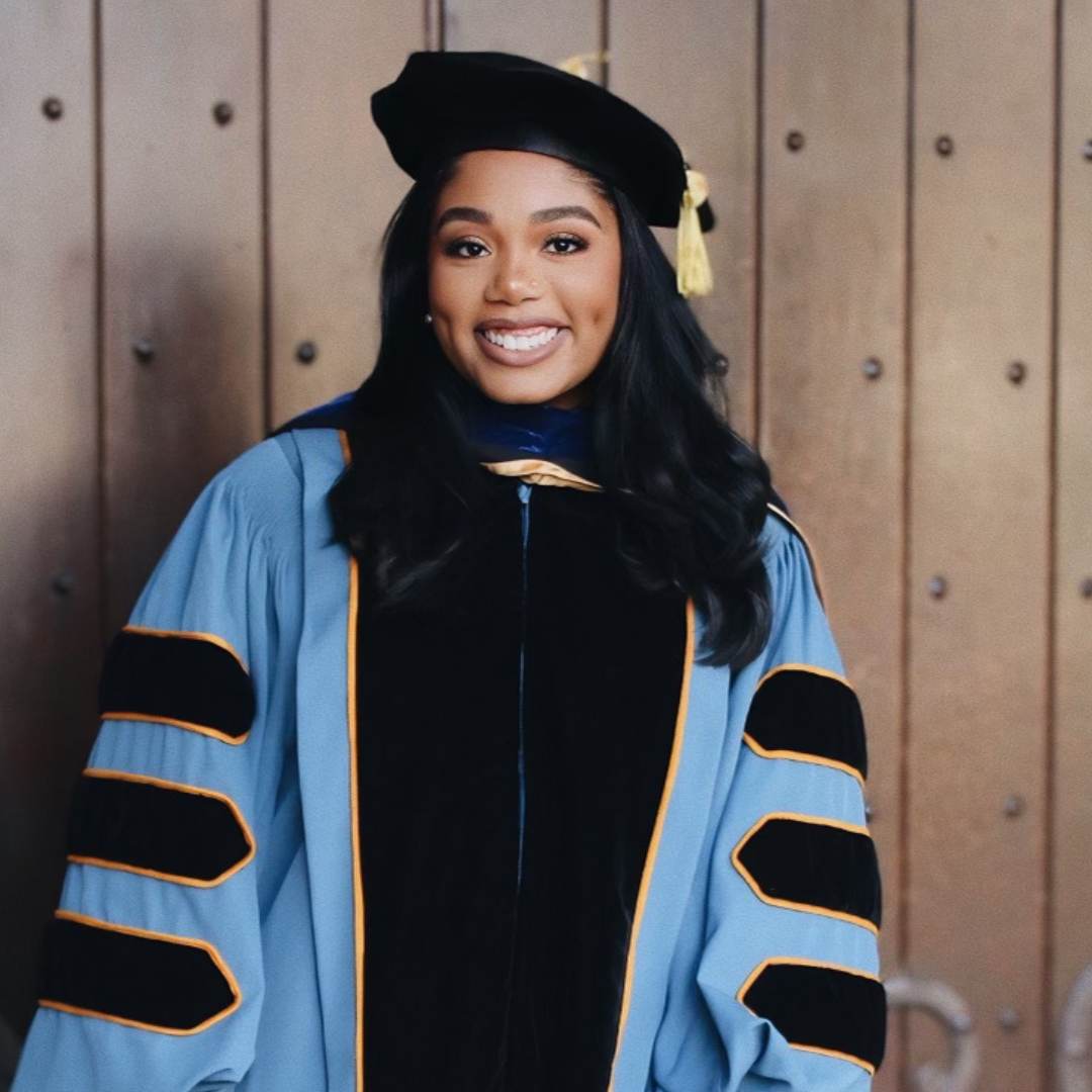 A woman in academic regalia with a doctoral gown and cap stands and smiles in front of a wooden door.