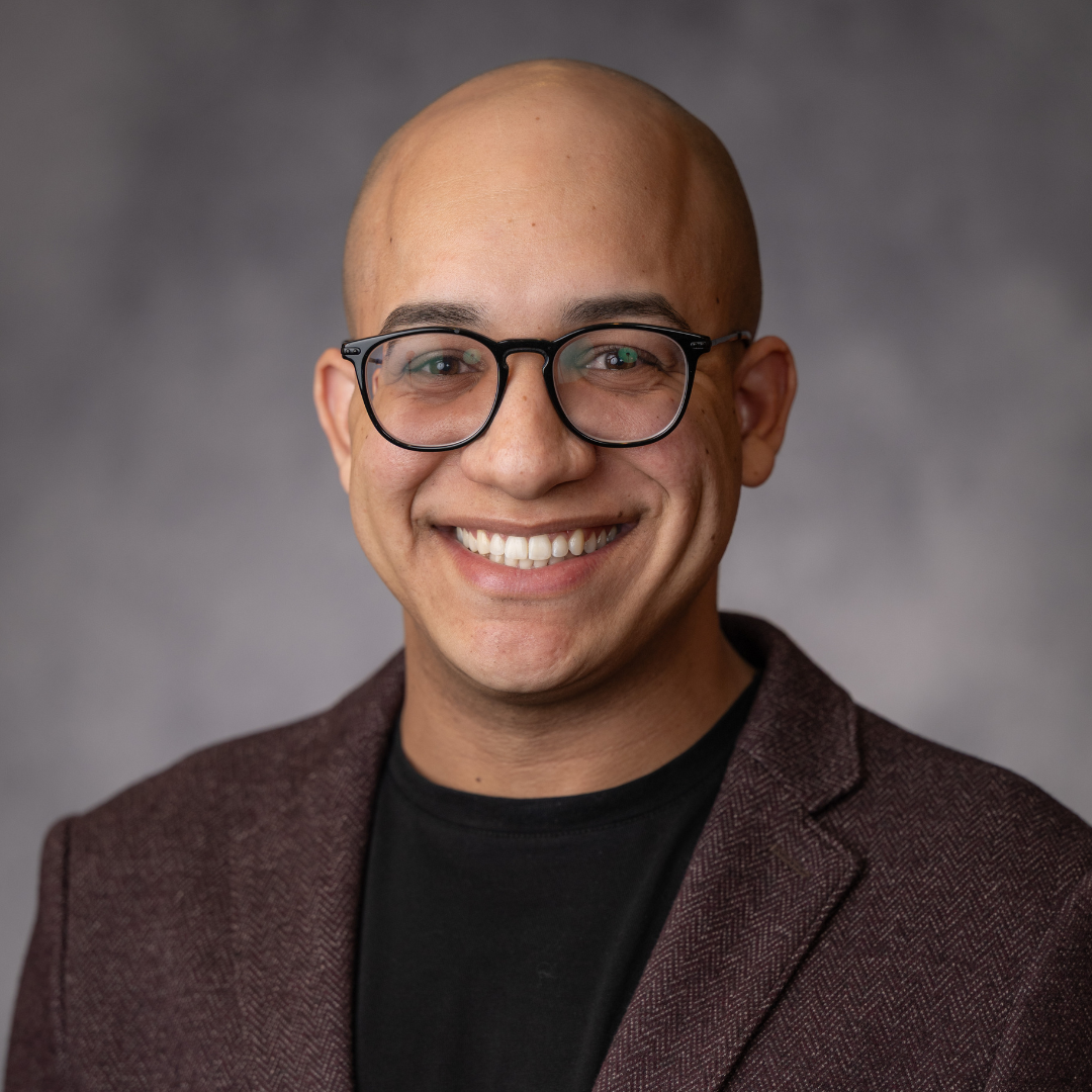 A person with glasses and a shaved head is smiling, wearing a dark jacket and black shirt, posing in front of a gray blurred background.