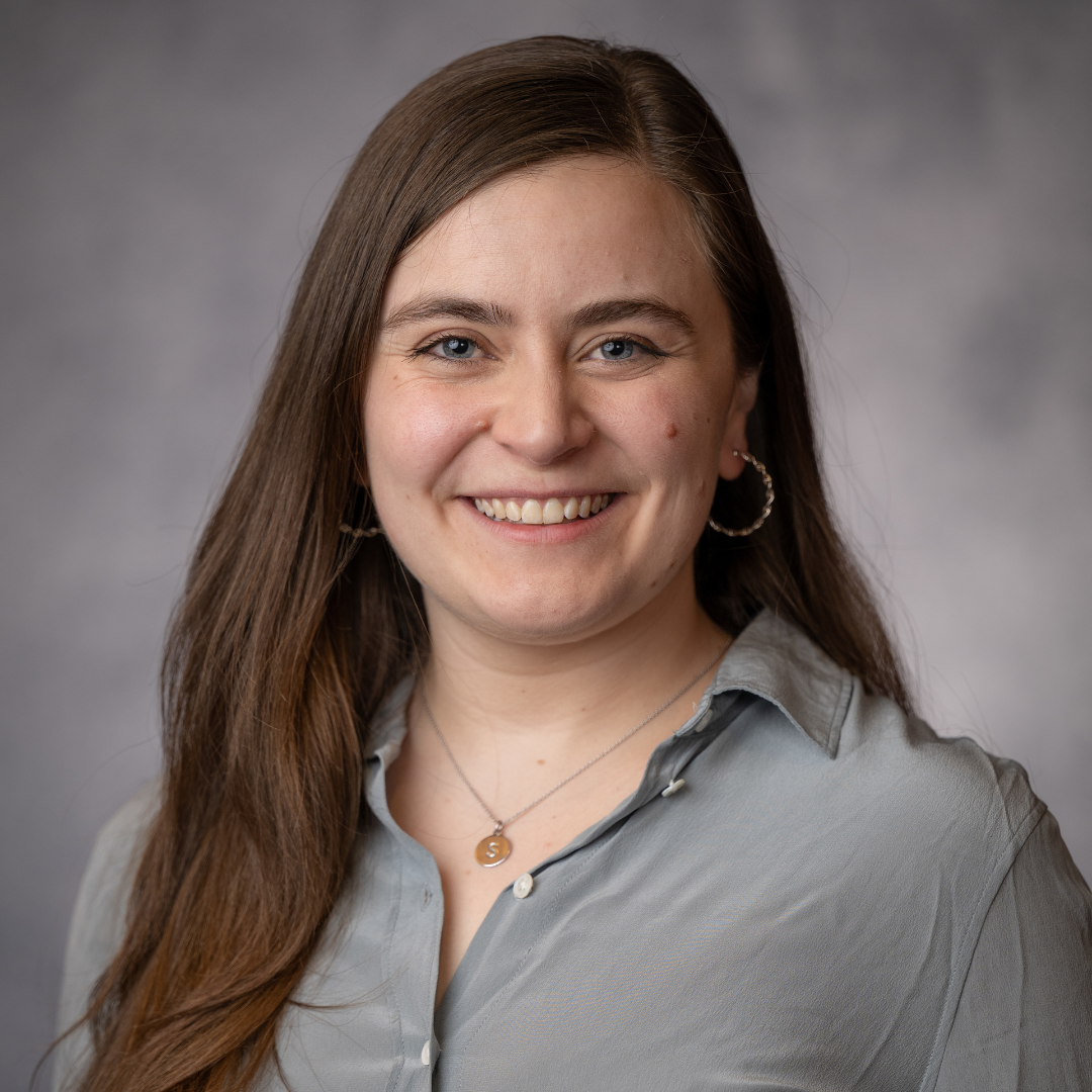 A woman with long brown hair, wearing a light gray blouse and hoop earrings, smiles at the camera against a neutral gray background.