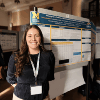 A woman stands in front of a scientific research poster about phenotypes at a conference, wearing a lanyard and smiling at the camera.