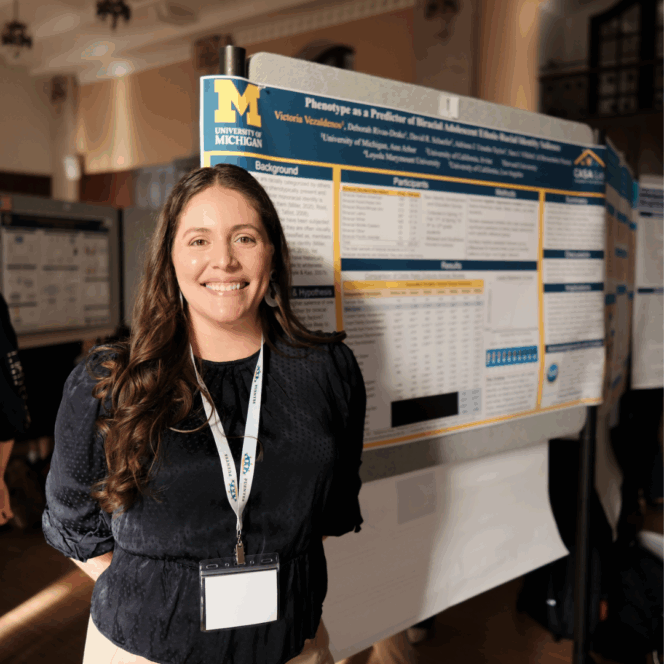 A woman stands in front of a scientific research poster about phenotypes at a conference, wearing a lanyard and smiling at the camera.