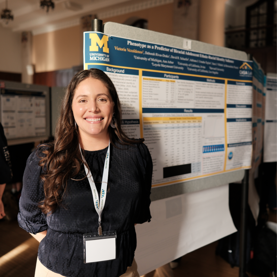 A woman stands in front of a scientific research poster about phenotypes at a conference, wearing a lanyard and smiling at the camera.