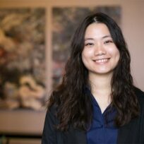 A woman with long wavy hair wearing a dark blazer and blue shirt smiles at the camera, standing indoors with blurred artwork in the background.