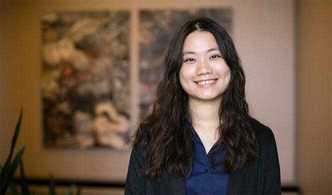 A woman with long wavy hair wearing a dark blazer and blue shirt smiles at the camera, standing indoors with blurred artwork in the background.