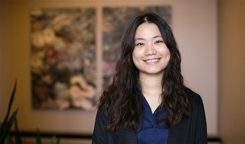 A woman with long wavy hair wearing a dark blazer and blue shirt smiles at the camera, standing indoors with blurred artwork in the background.