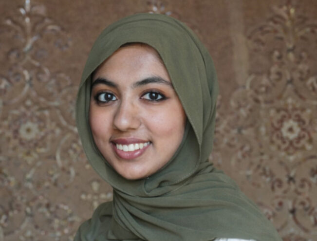 A woman wearing an olive green hijab and white top smiles at the camera, standing in front of a patterned brown background.