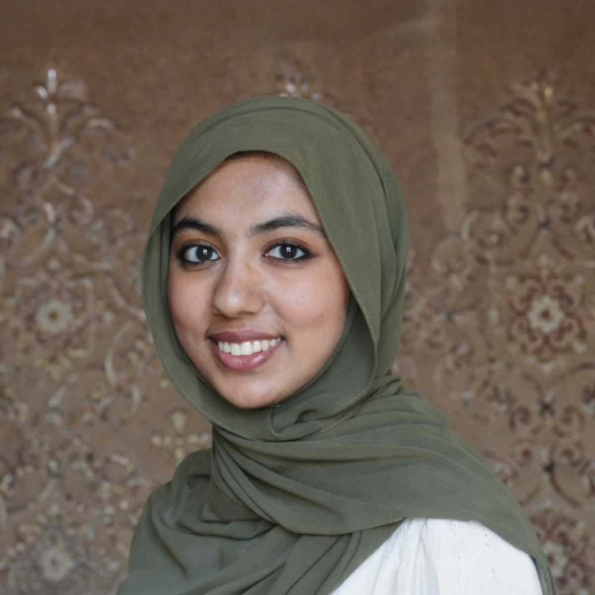 A woman wearing an olive green hijab and white top smiles at the camera, standing in front of a patterned brown background.