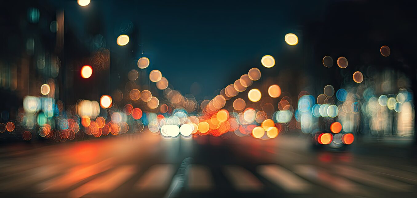 Blurred city street at night with out-of-focus car headlights, streetlights, and traffic signals creating colorful bokeh effects.