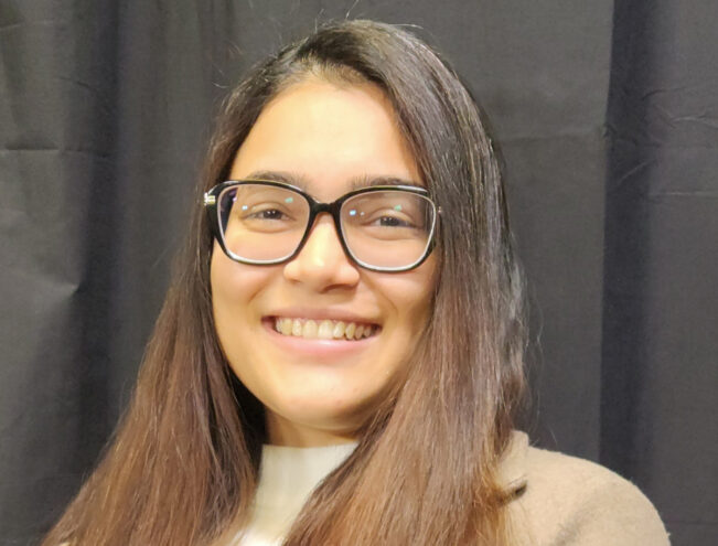 A woman with long brown hair and glasses smiles at the camera, standing in front of a black curtain while wearing a beige coat and white top.