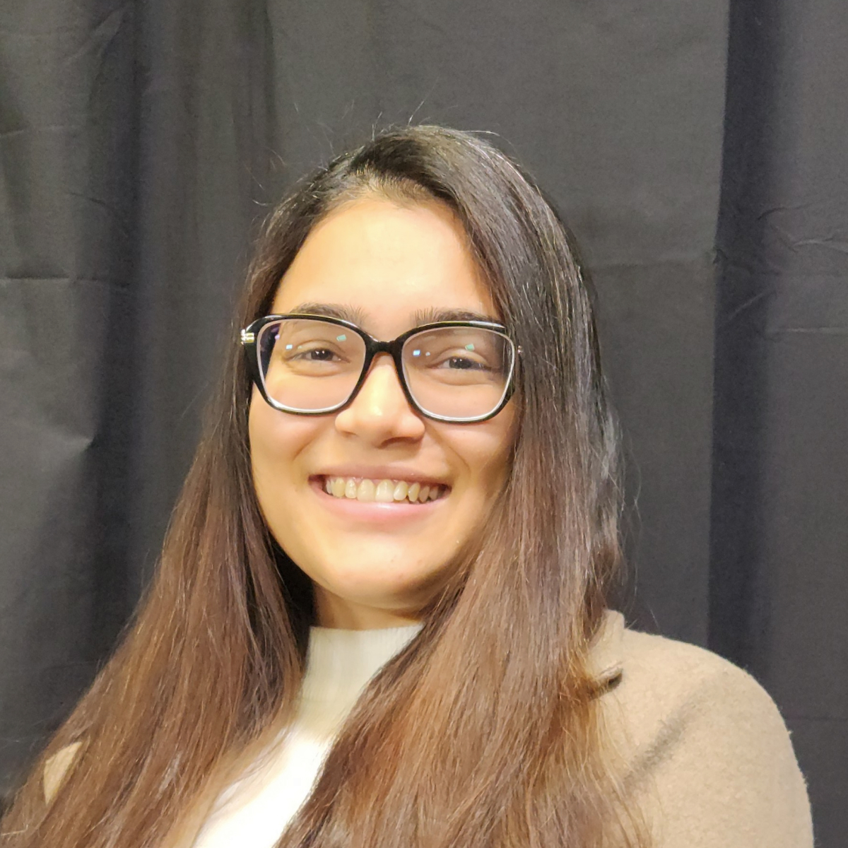 A woman with long brown hair and glasses smiles at the camera, standing in front of a black curtain while wearing a beige coat and white top.