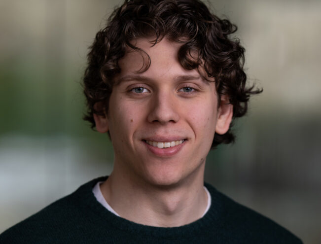 A young man with curly brown hair and a dark green sweater smiles at the camera against a blurred background.